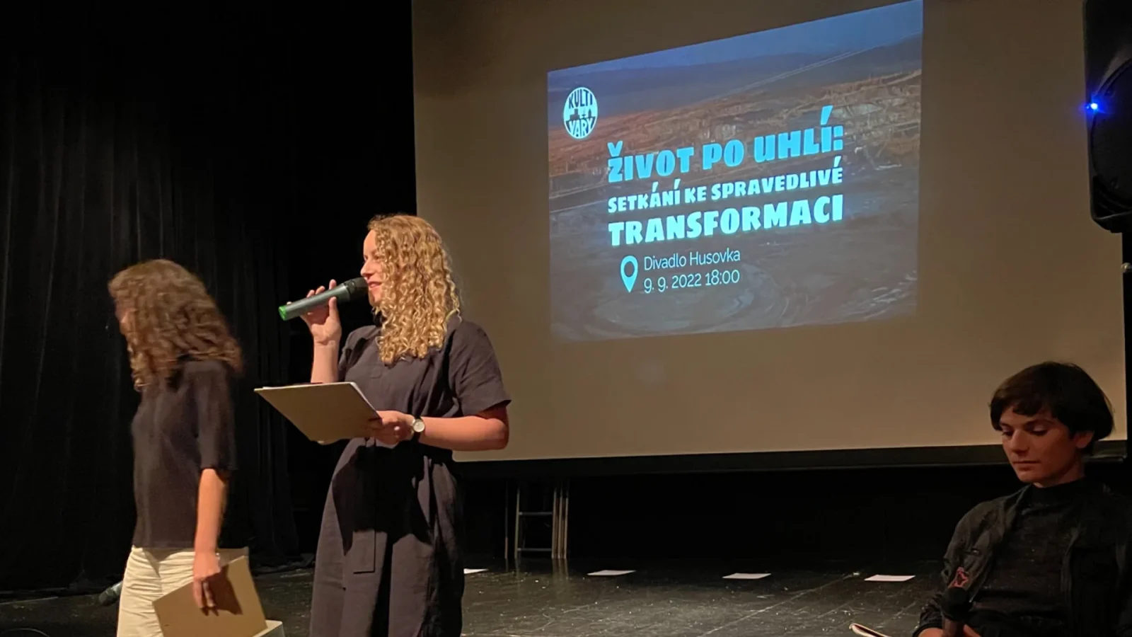 Veřejné setkání Život po uhlí, moderátorka s mikrofonem před promítanou pozvánkou.